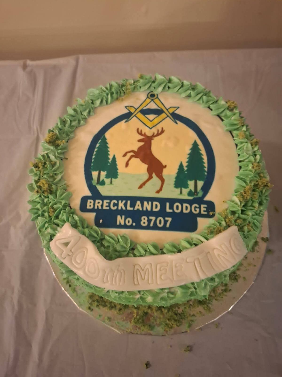 Breckland Lodge No. 8707 cake decorated with green icing, featuring the lodge emblem with a rearing deer, Masonic square and compass symbol, and trees.