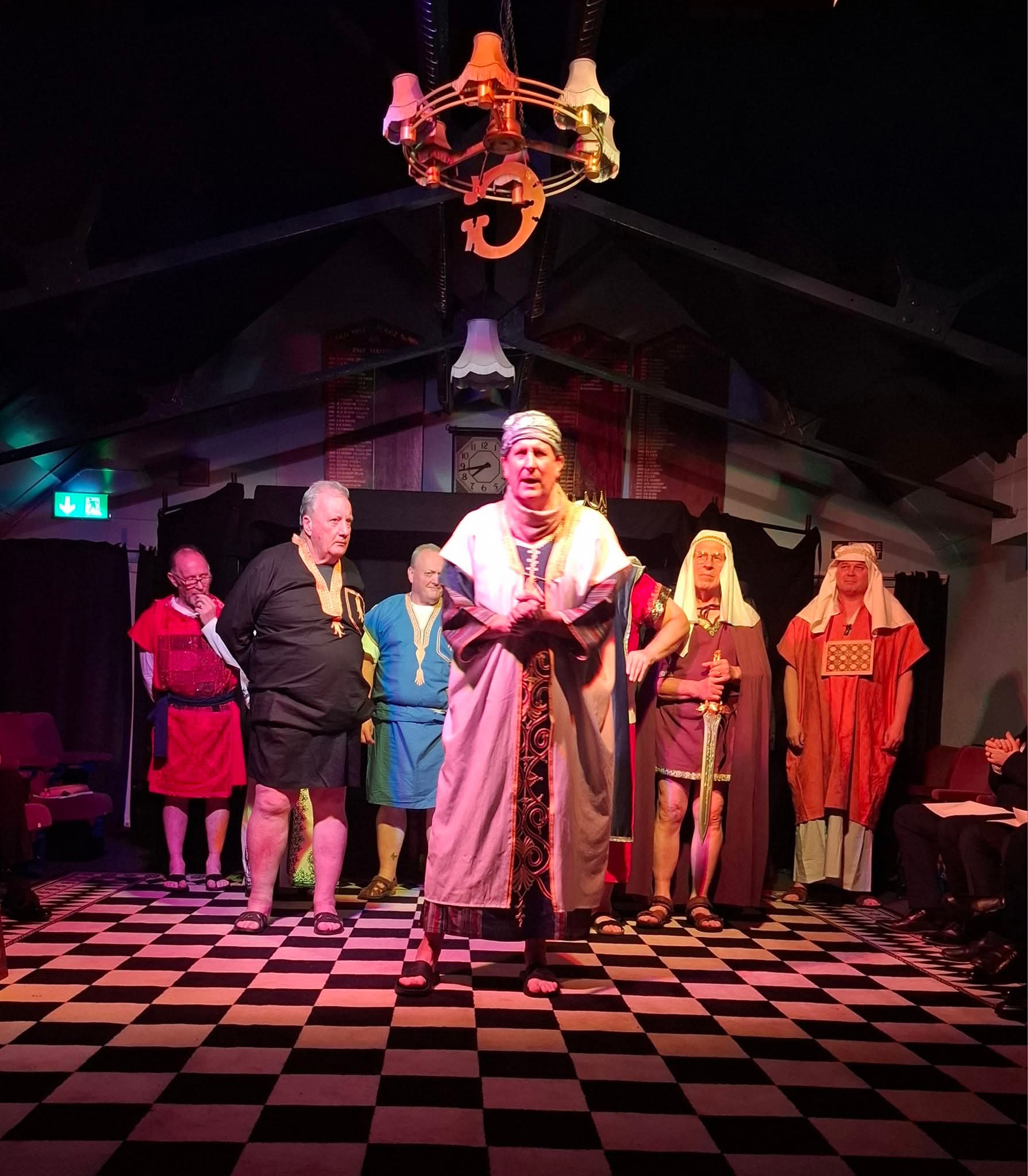 Masonic lodge members in ceremonial regalia standing on a checkered floor beneath illuminated symbols.